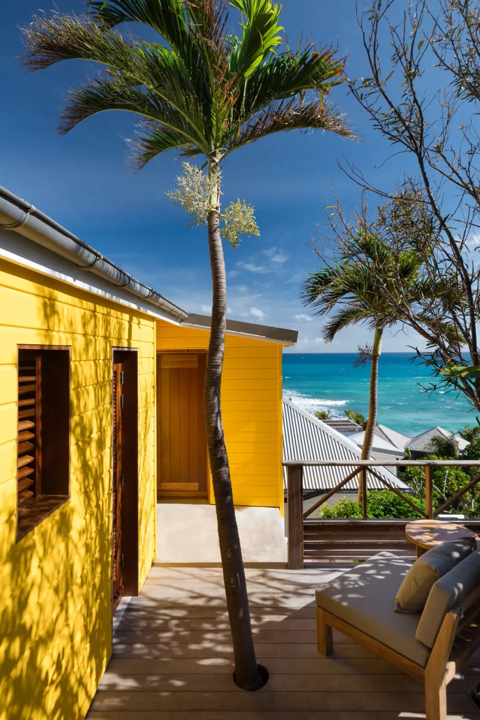 Ocean View room deck at Hotel Manapany beside a yellow building with palms and sea beyond.