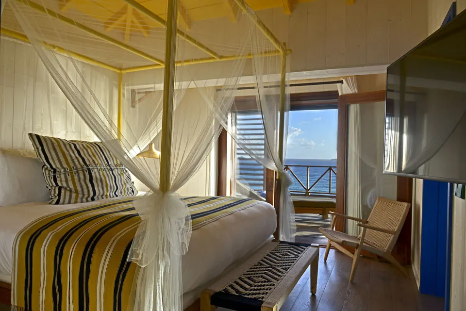 Ocean View room at Hotel Manapany with a mosquito-net bed, chair, TV and balcony sea view.