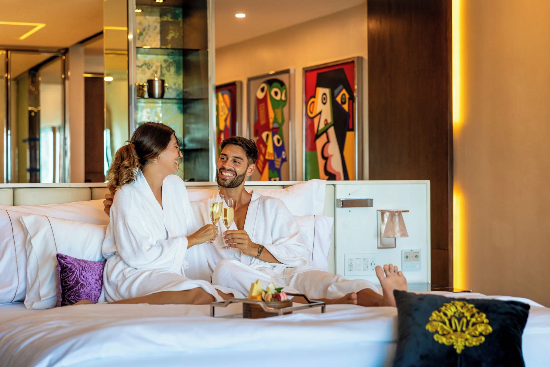 Couple in robes toasting in a guest room bed at Hotel Mousai Cancun.