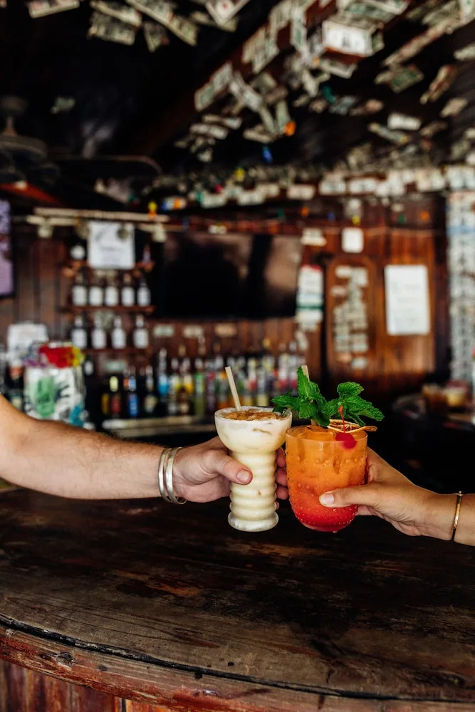 Two cocktails held over a wooden bar inside Beach Bar at Hotel on the Cay.
