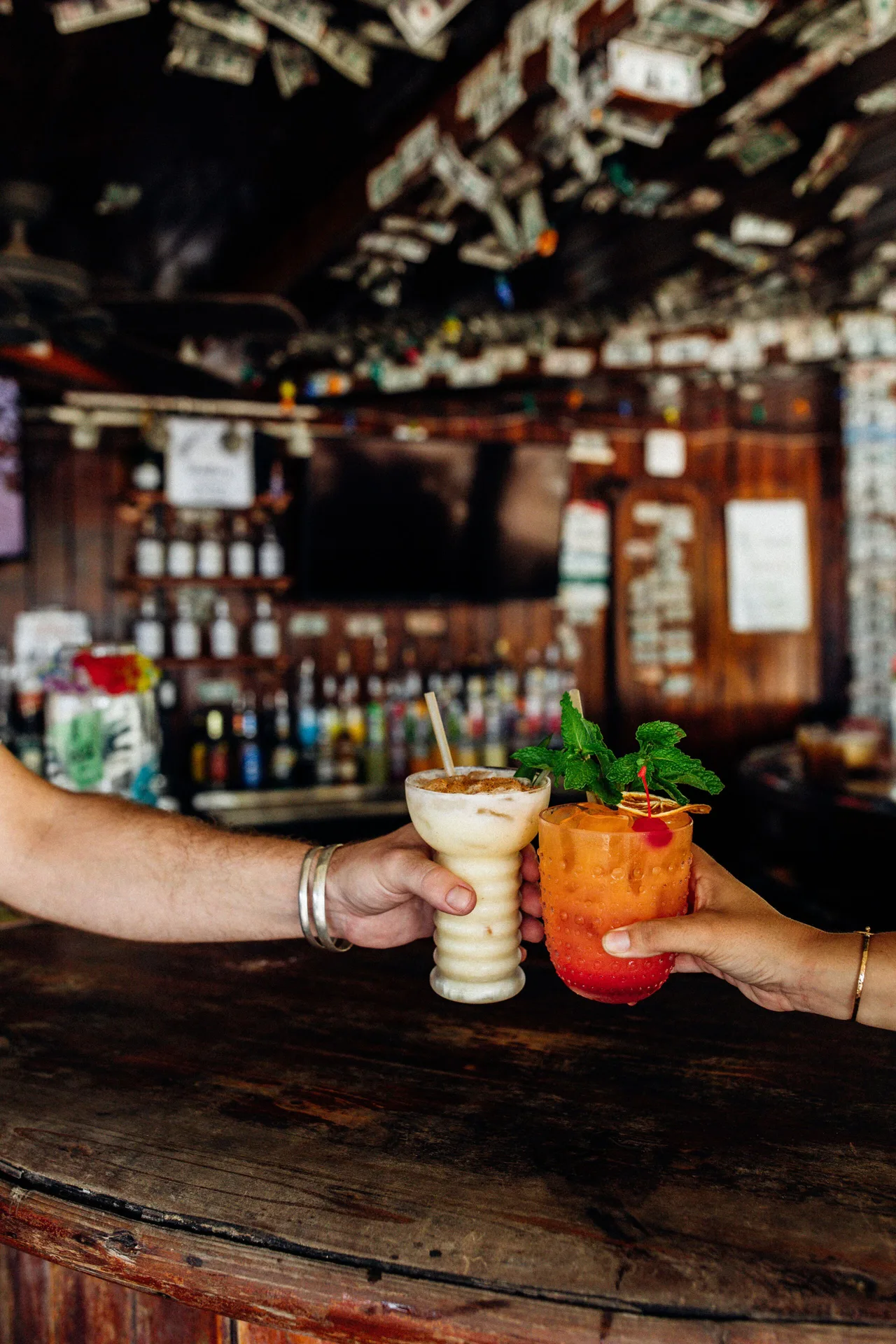 Two cocktails being raised over a dark wooden bar at Hotel on the Cay.
