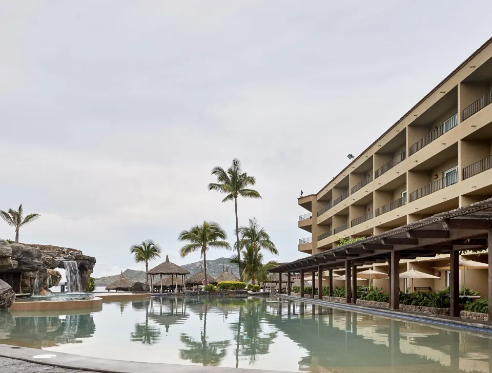 Lagoon-style pool with waterfall, palms and ocean beyond at Hotel Playa Mazatlan.