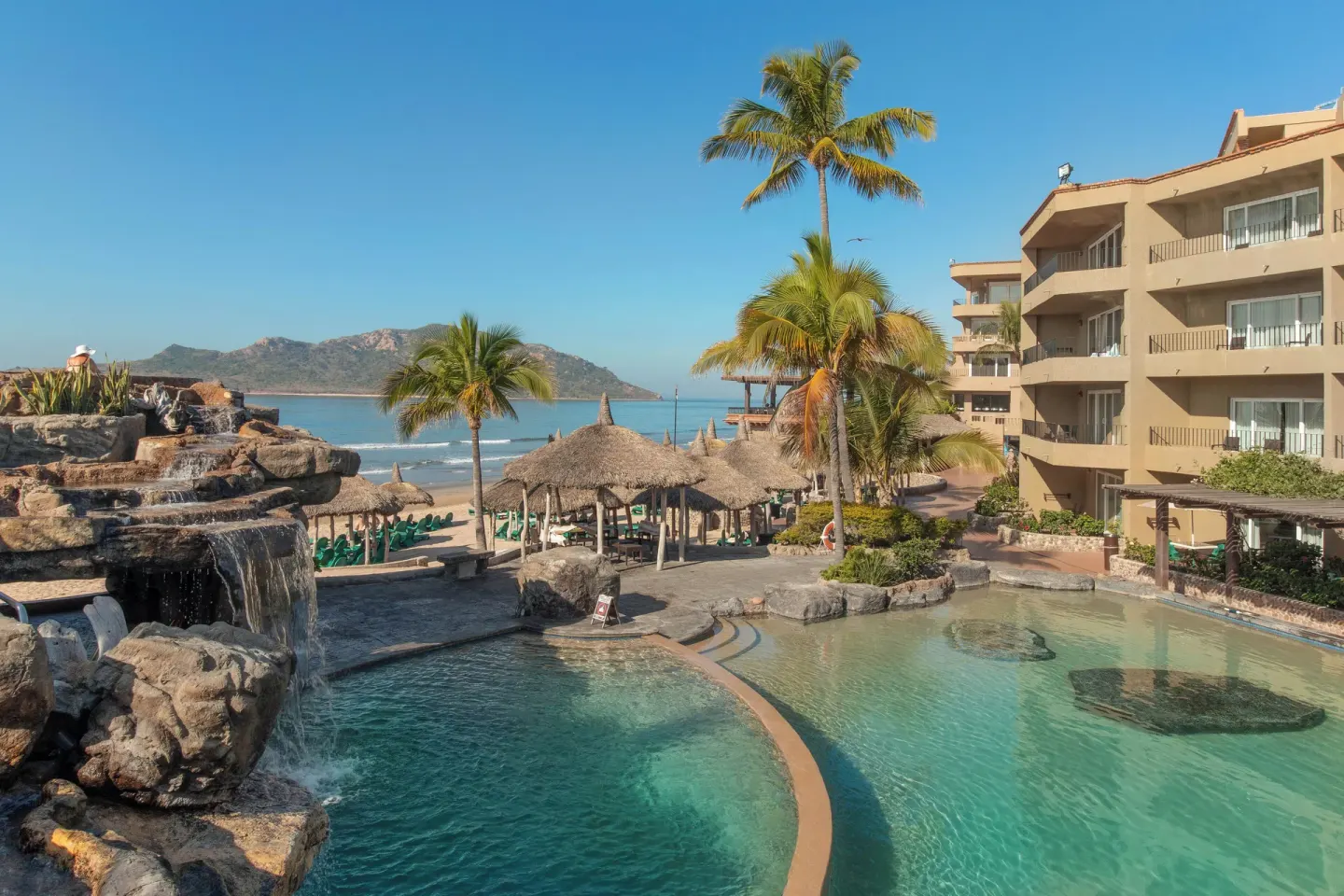 Oceanfront pool with waterfall, palms, palapas, and beach beyond at Hotel Playa Mazatlan.