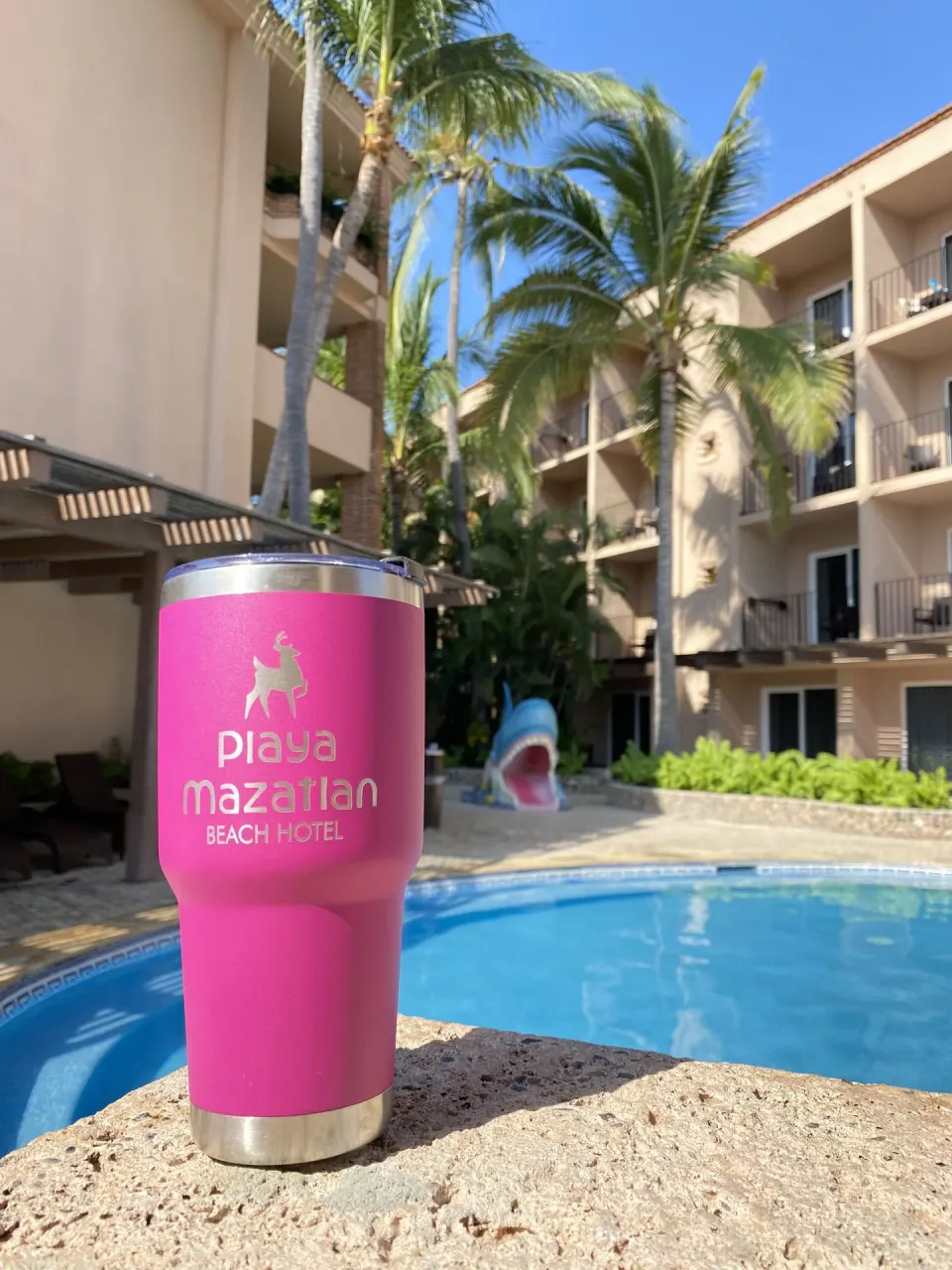 Pink Hotel Playa Mazatlan tumbler beside a pool with balconies and palms in the background.
