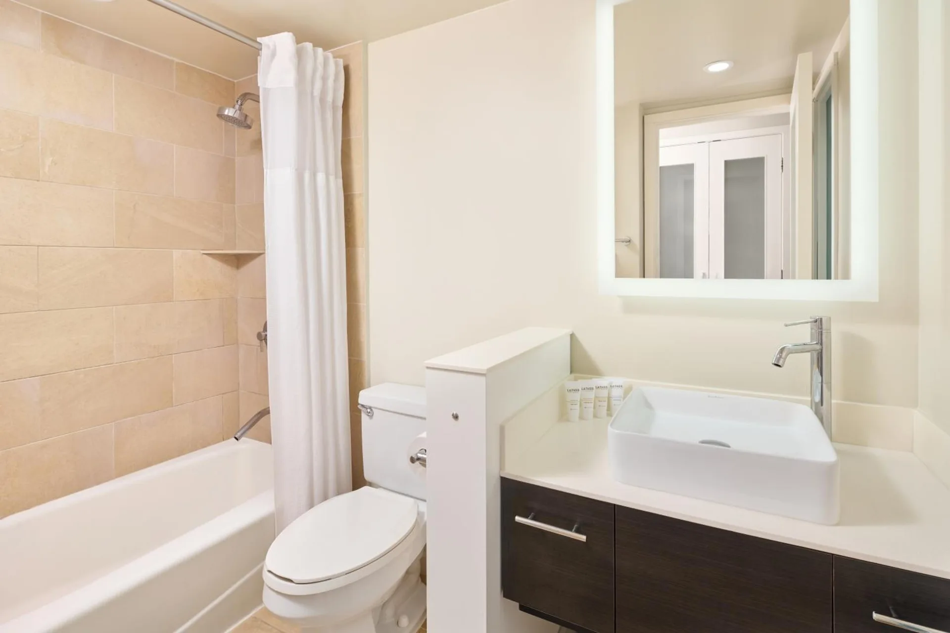 A bathroom with a tub shower, toilet, vessel sink, and lit mirror is shown at Hotel Renew.