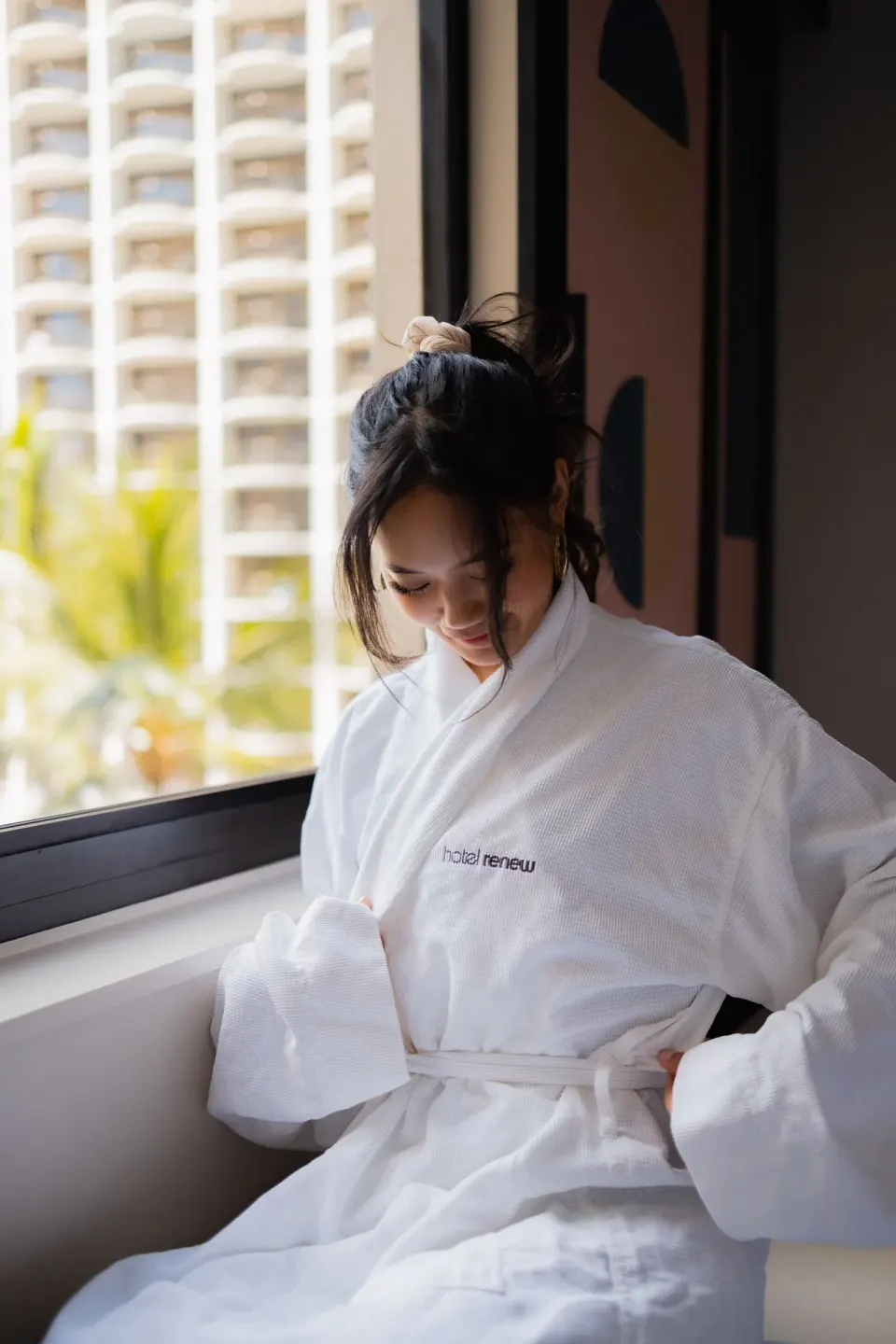 A guest wears a white Hotel Renew robe while sitting by a window.