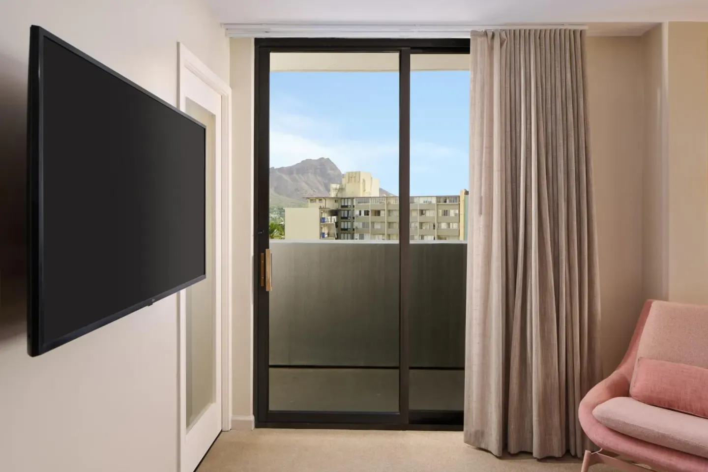 A guest room TV, pink chair, and balcony door with a mountain view appear at Hotel Renew.