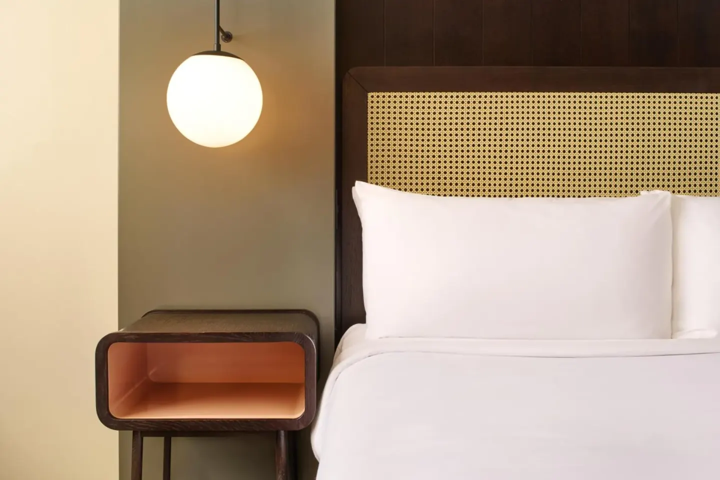 A white bed with a cane headboard, bedside table, and globe lamp is shown at Hotel Renew.