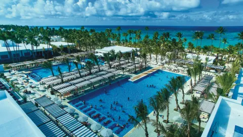 High view over the main pools, shade pergolas, palms, and ocean at Hotel Riu Bambu.