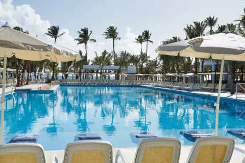 Pool view from loungers under umbrellas with palms around Hotel Riu Bambu.