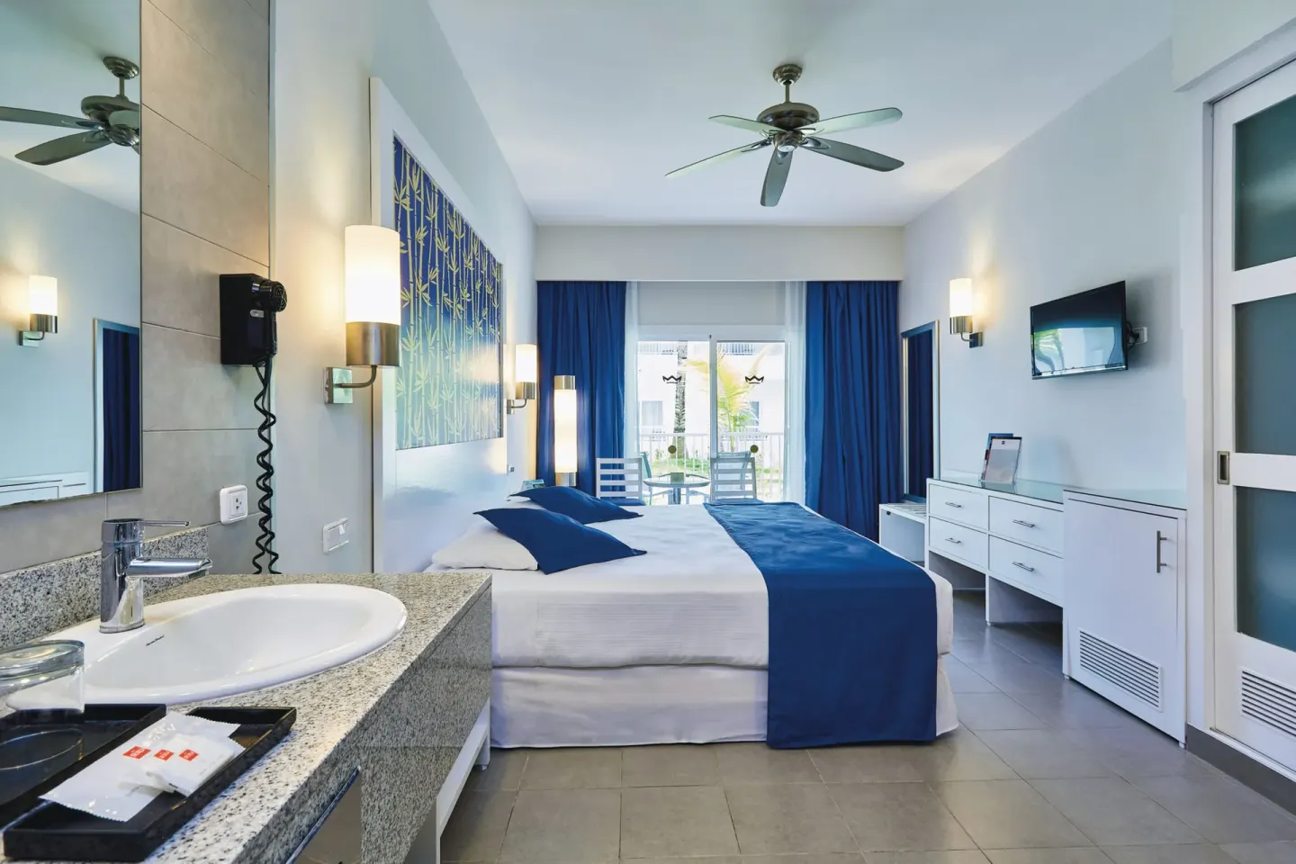 Deluxe Room with bed, balcony doors, dresser, TV, and vanity at Hotel Riu Bambu.