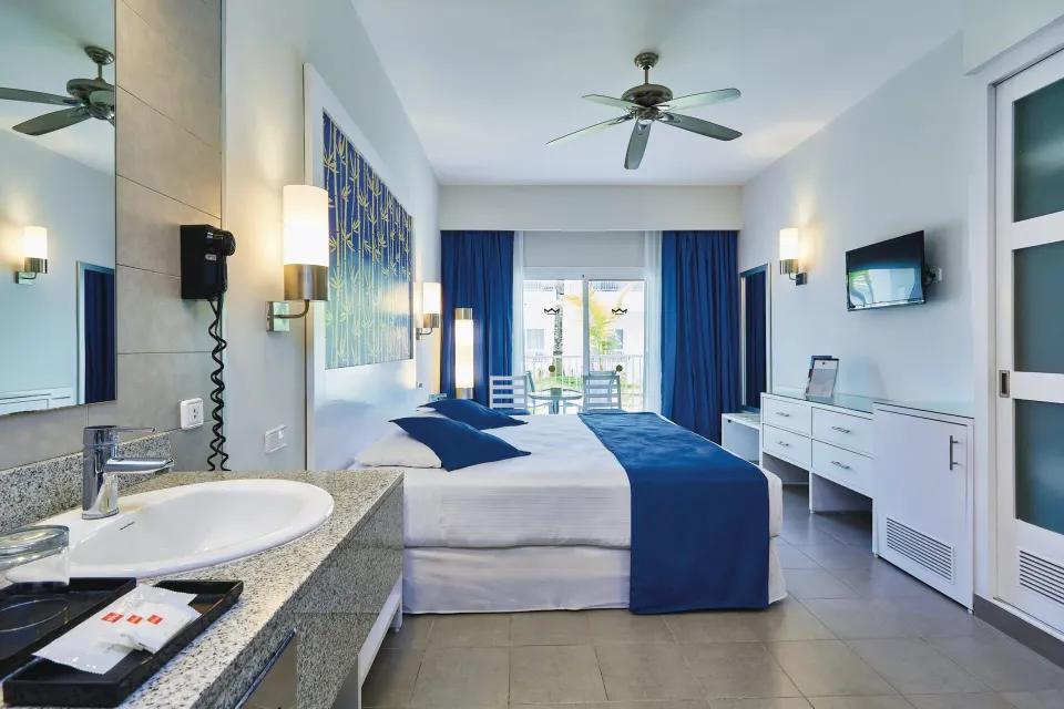 Deluxe Room with bed, balcony doors, dresser, TV, and vanity at Hotel Riu Bambu.