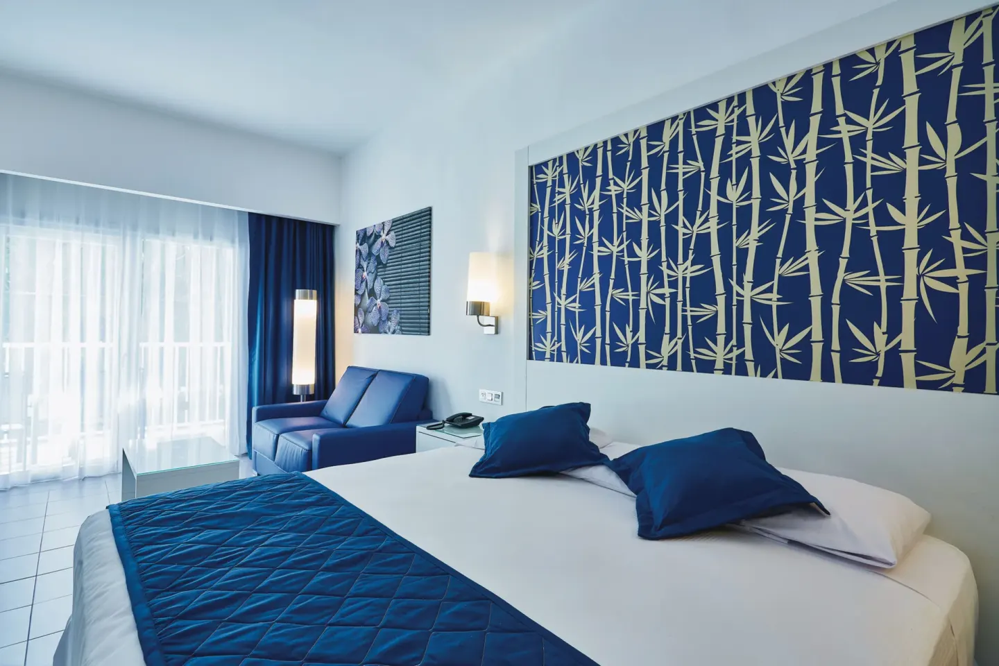 Deluxe Room bed with blue sofa and balcony doors at Hotel Riu Bambu.