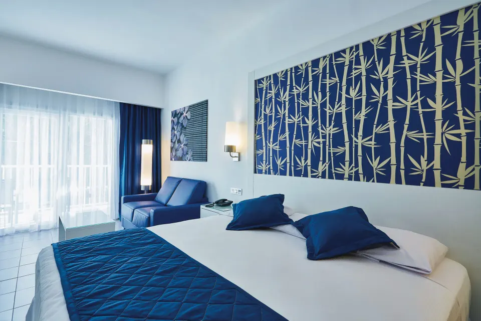 Deluxe Room bed with blue sofa and balcony doors at Hotel Riu Bambu.