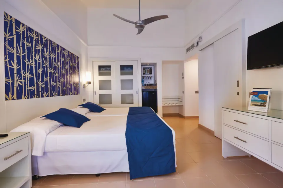 Guest room with a king bed, blue bedding, dresser, TV and beverage area at Hotel Riu Bambu.