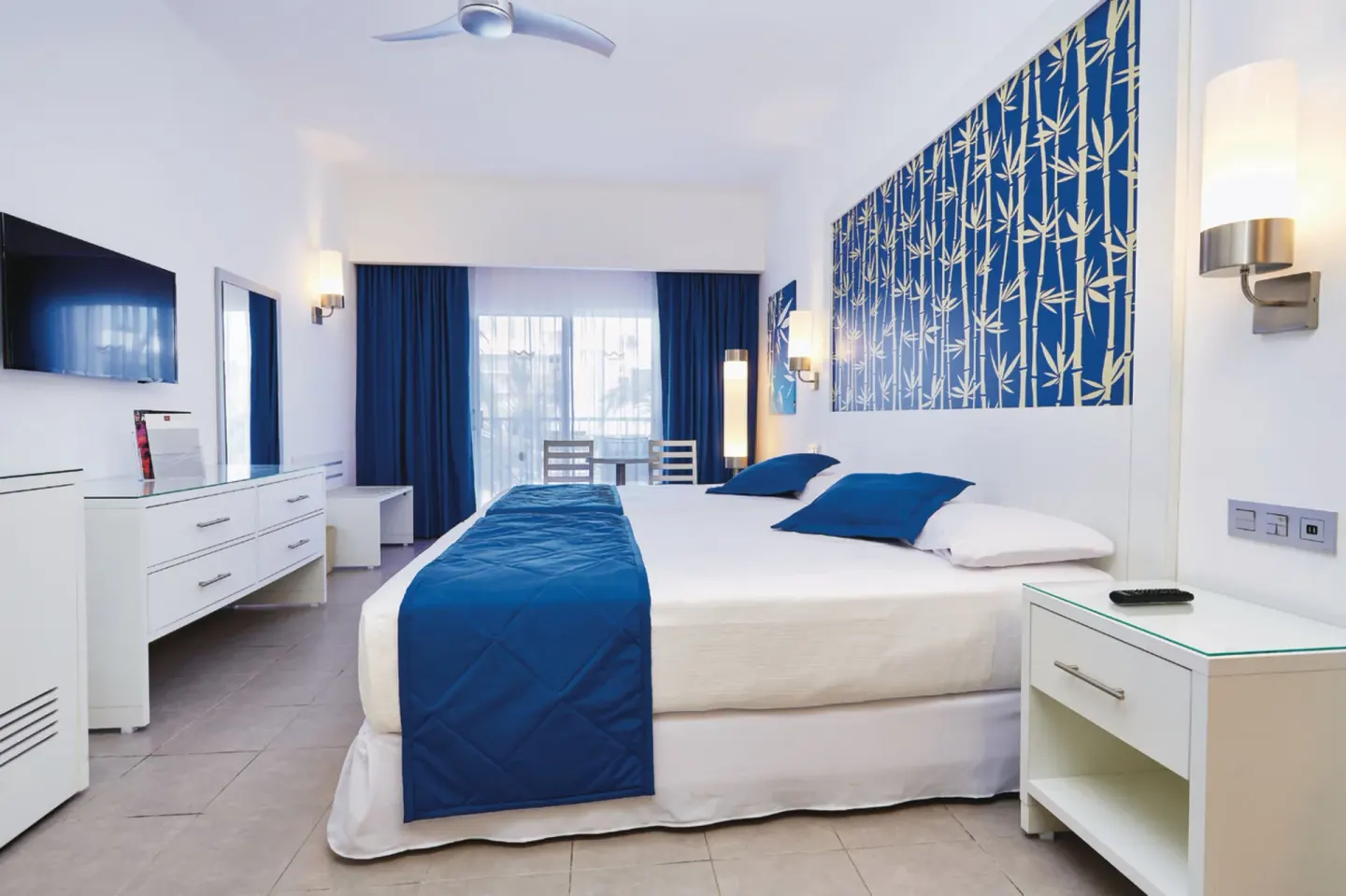 Guest room with a king bed, blue accents, wall TV and balcony doors at Hotel Riu Bambu.