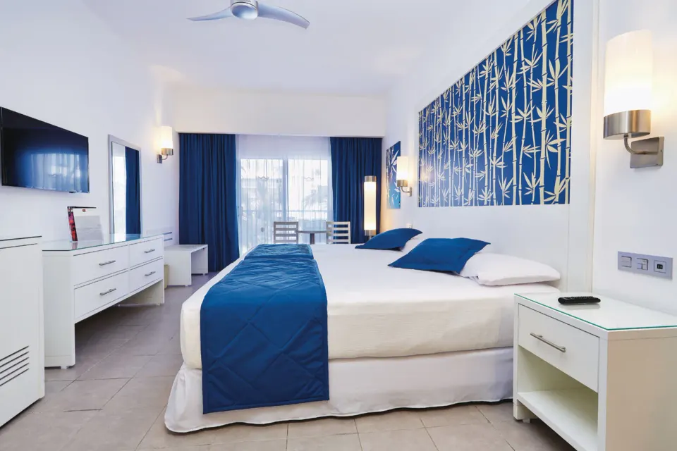 Guest room with a king bed, blue accents, wall TV and balcony doors at Hotel Riu Bambu.