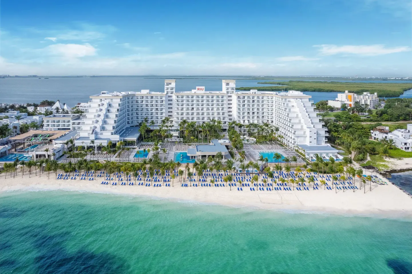 Aerial beachfront view of Hotel Riu Caribe with pools, palms, sand, and blue-green water.