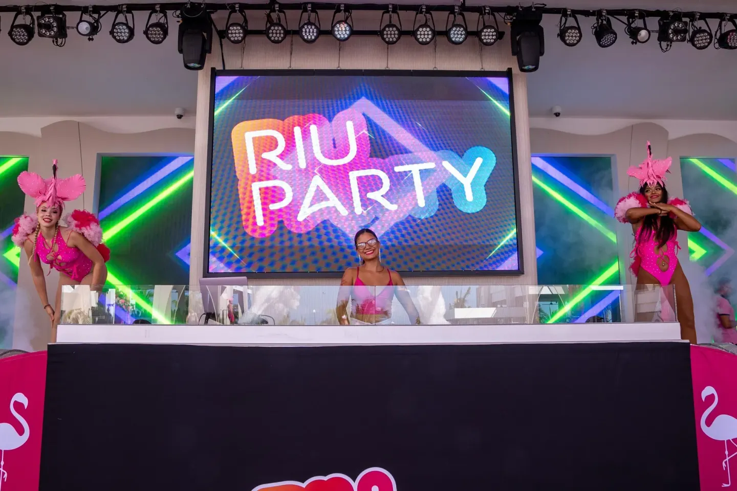 DJ booth and dancers stand before a bright party screen at Hotel Riu Caribe.