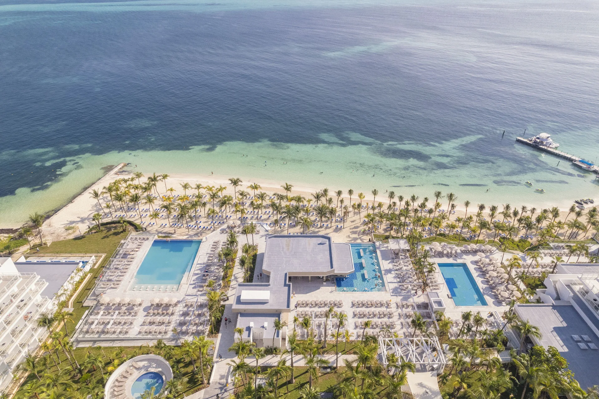 Overhead view of pools, palm-lined beach, turquoise shallows, and pier at Hotel Riu Caribe.