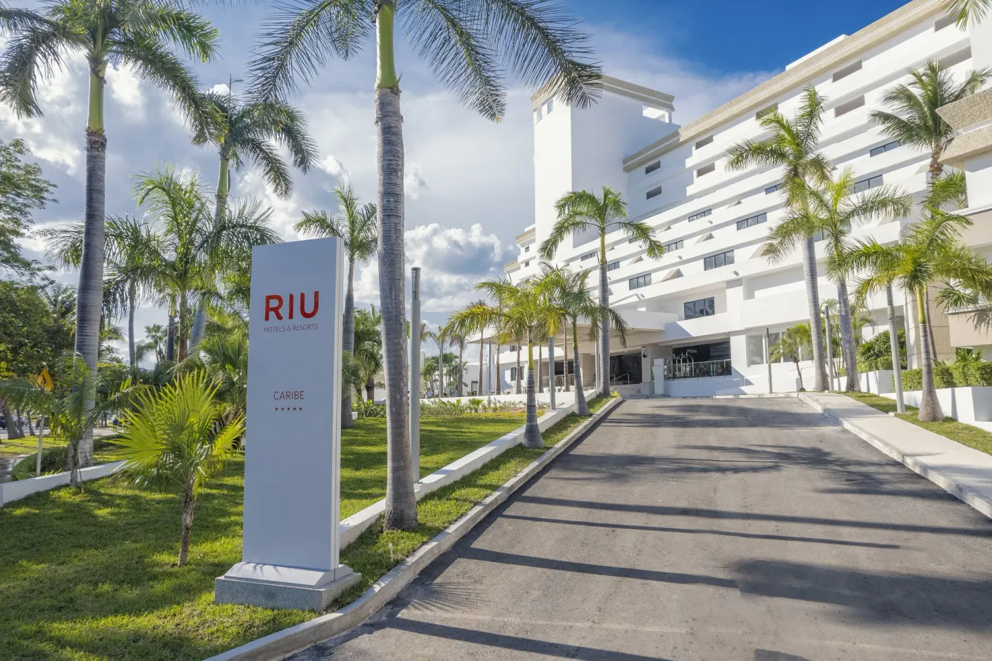 Hotel Riu Caribe entrance sign beside a palm-lined driveway and white building.