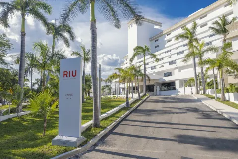 Hotel Riu Caribe entrance sign beside a palm-lined driveway and white building.