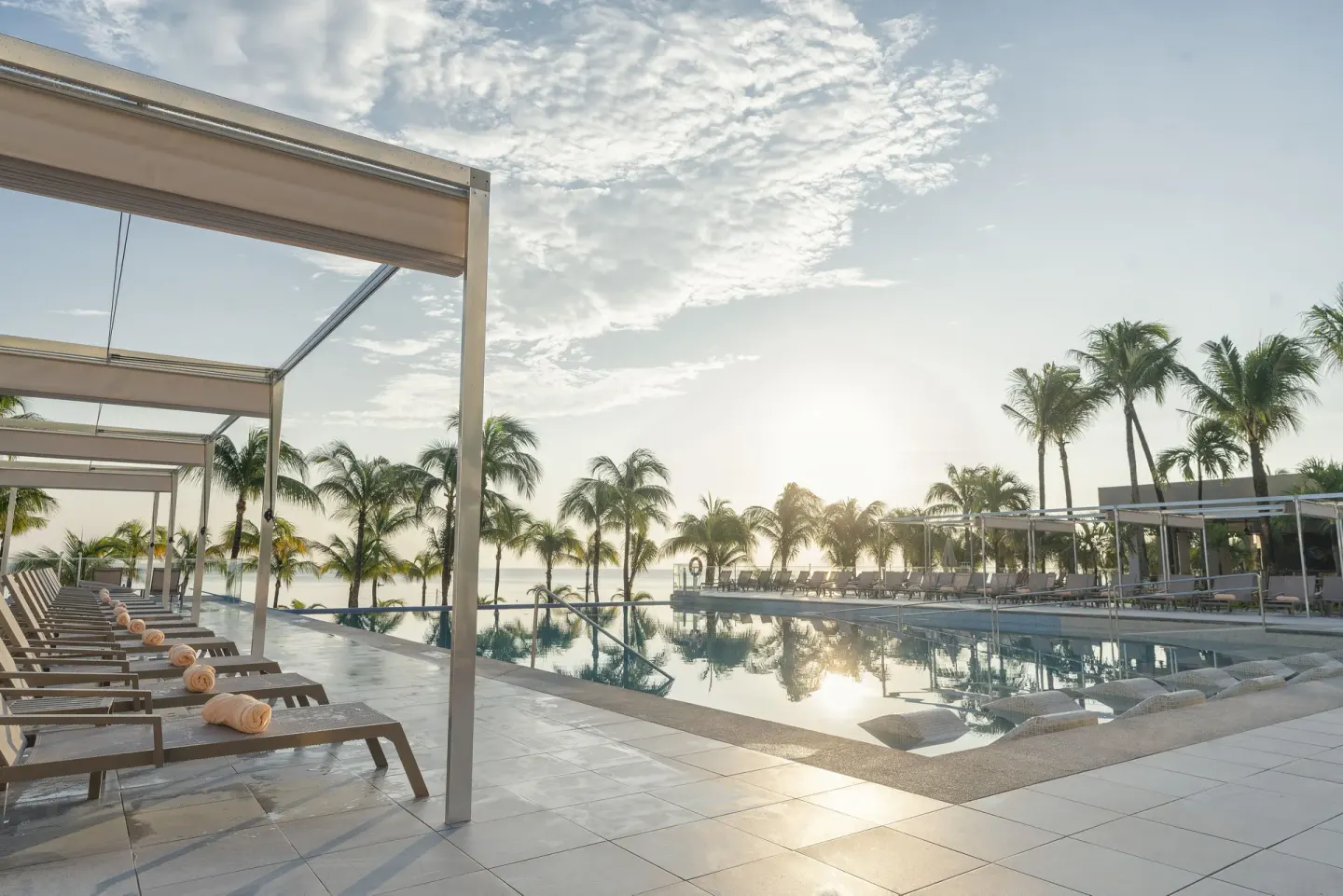 Pool deck with loungers, shade structures, palms, and ocean light at Hotel Riu Caribe.