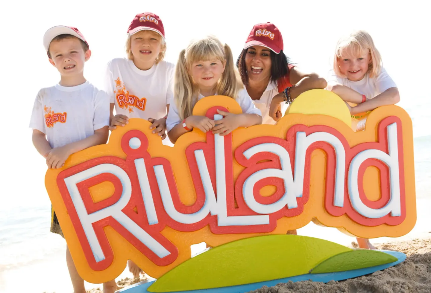 Children and a staff member pose with a RiuLand sign on the beach at Hotel Riu Caribe.