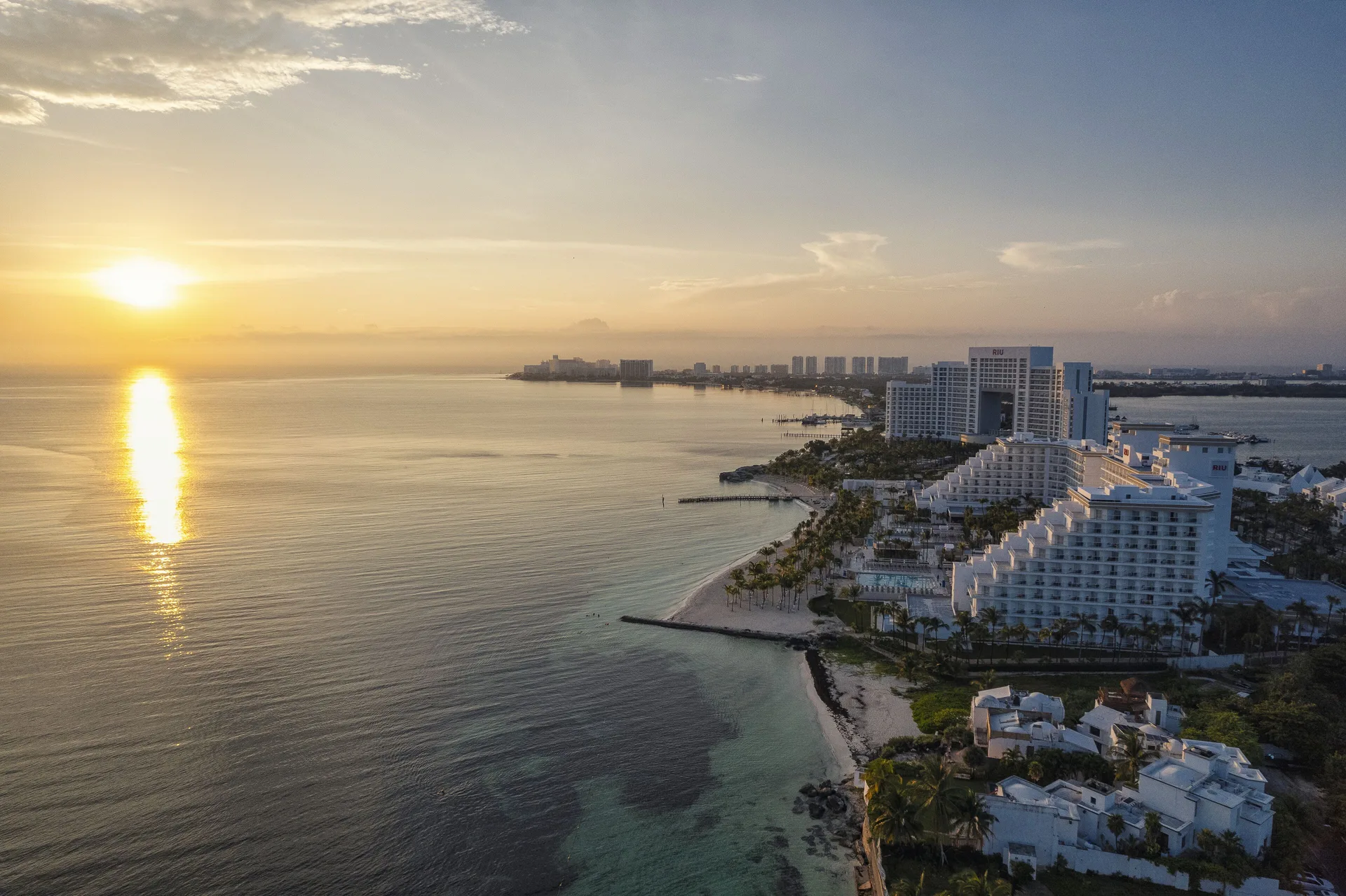 Sunset coastline view with Hotel Riu Caribe beside calm water and nearby buildings.