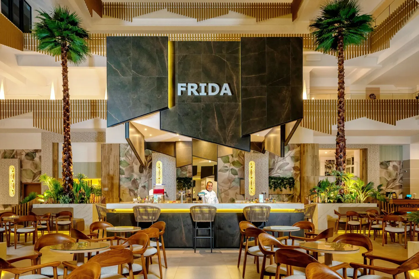 Frida lobby bar with palm trees, round tables and a central counter at Hotel Riu Caribe.