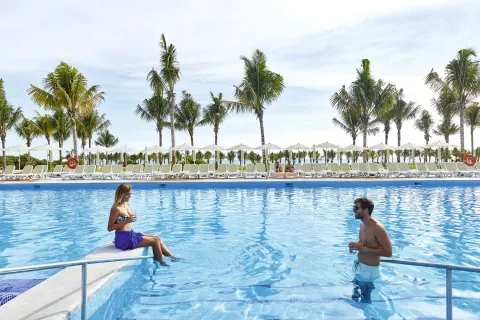 Guests stand and sit at the edge of a large outdoor pool at Hotel Riu Dunamar.