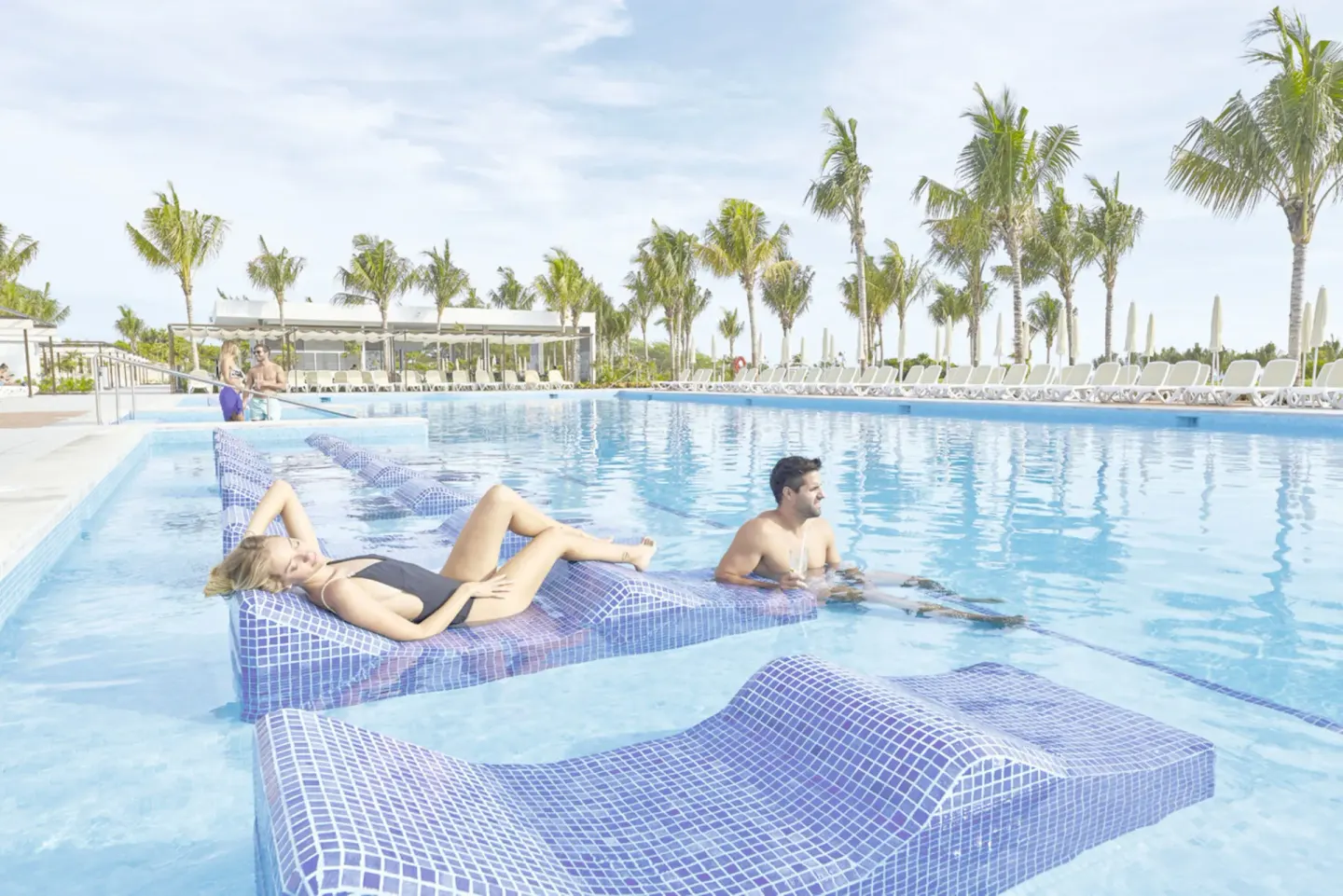 Guests relax on submerged loungers in a palm-lined pool at Hotel Riu Dunamar.