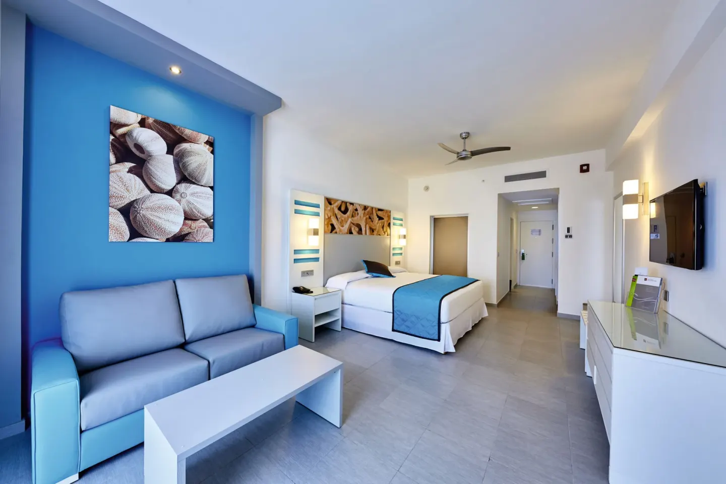 Oceanfront standard room with king bed, blue sofa, coffee table, and TV at Hotel Riu Dunamar.