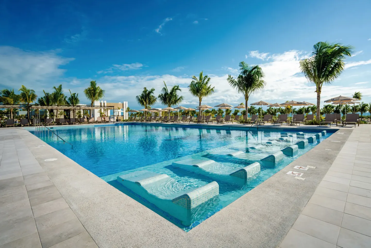 Corner view of a pool with submerged loungers and palm-lined seating at Hotel Riu Latino.