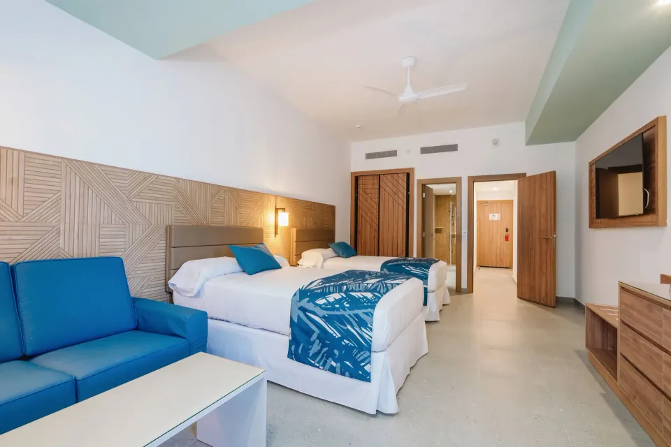 Guest room with two beds, blue sofa, TV, and open bathroom area at Hotel Riu Latino.