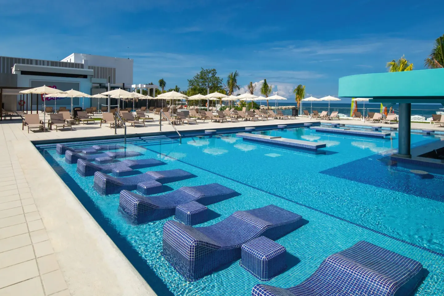 Pool with blue tiled loungers, umbrellas, and ocean beyond at Hotel Riu Montego Bay.