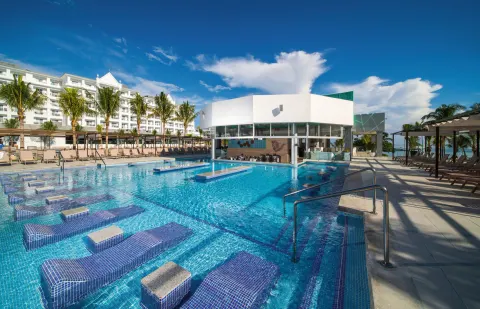 Main pool with submerged loungers, bar building and sea beyond at Hotel Riu Ocho Rios.