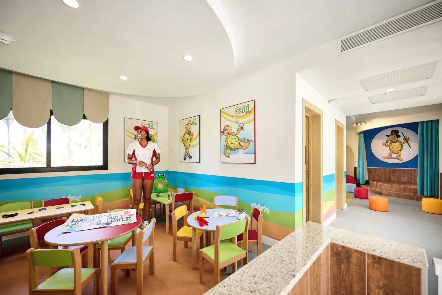 Children's activity room with small tables and stage at Hotel Riu Palace Aquarelle.