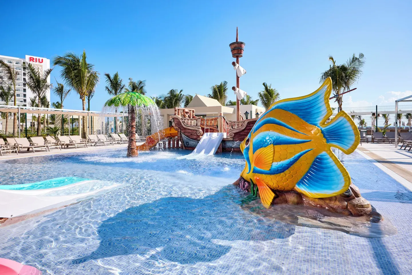 Kids splash pool with fish sculpture, pirate ship, slides, and water spray at Hotel Riu Palace Aquarelle.