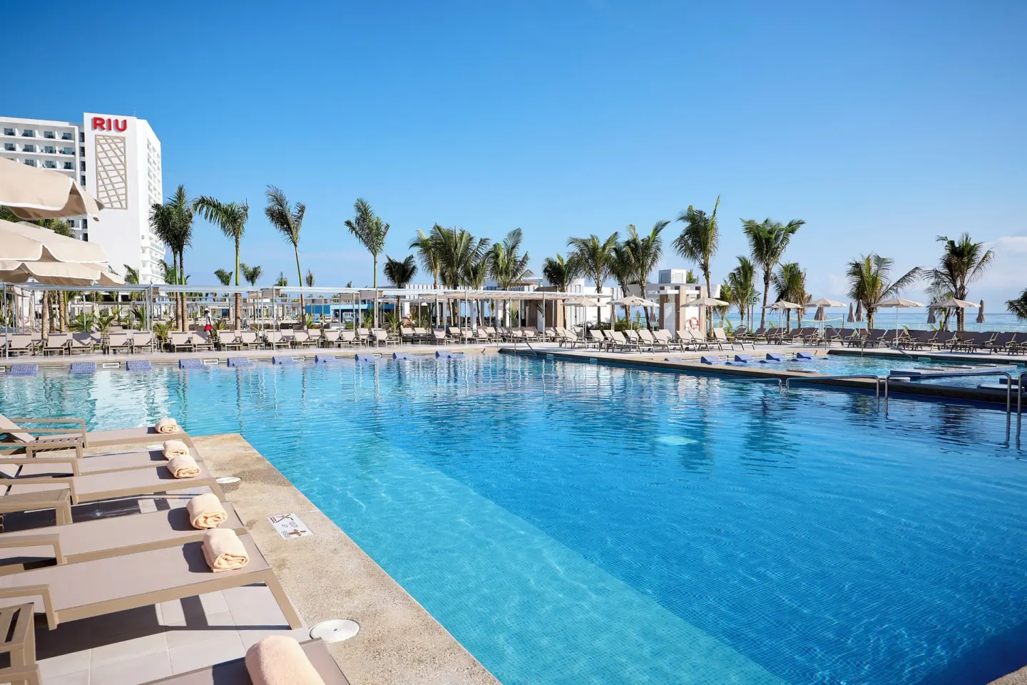Large pool with loungers, towels, palms, and ocean backdrop at Hotel Riu Palace Aquarelle.