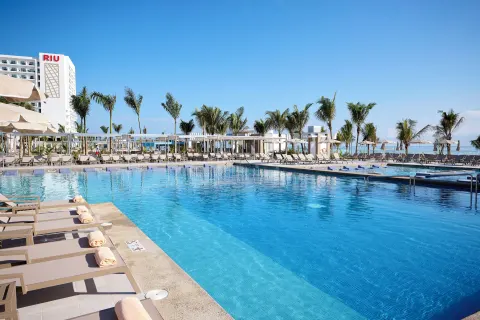 Large pool with loungers, towels, palms, and ocean backdrop at Hotel Riu Palace Aquarelle.