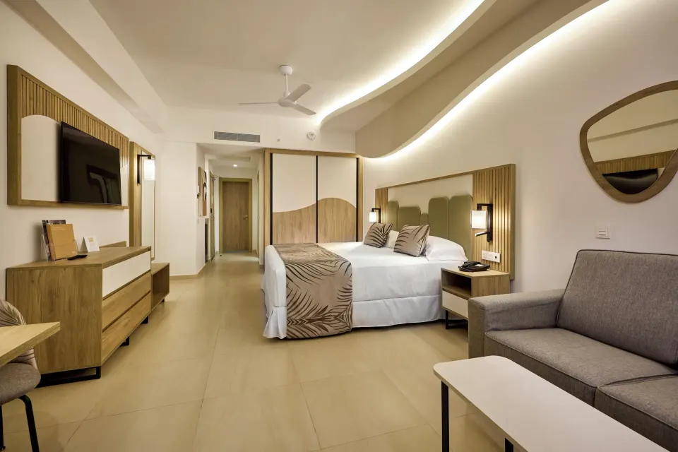 Junior Suite Partial Ocean View with bed, sofa, TV, and wardrobe at Hotel Riu Palace Aquarelle.