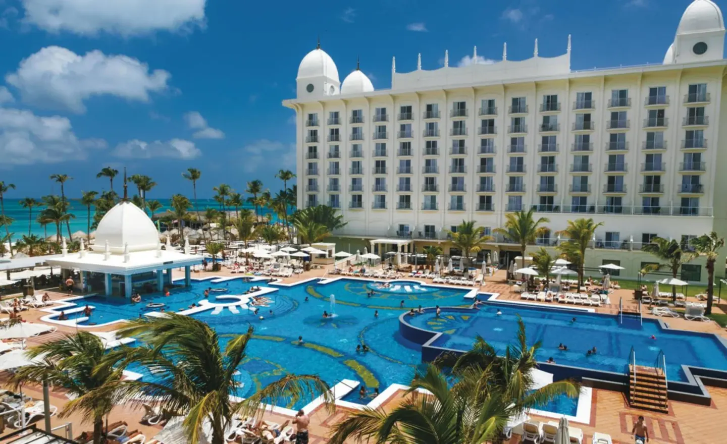 Pool, resort tower, palms, and ocean at Hotel Riu Palace Aruba