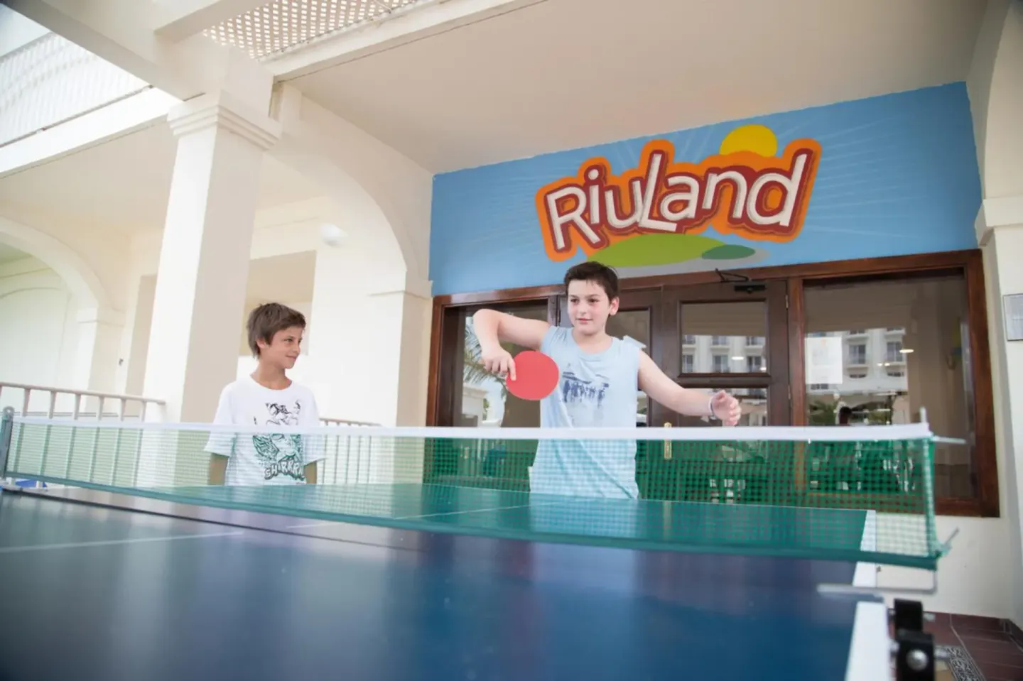 Two children play table tennis in a covered activity area at Hotel Riu Palace Aruba.