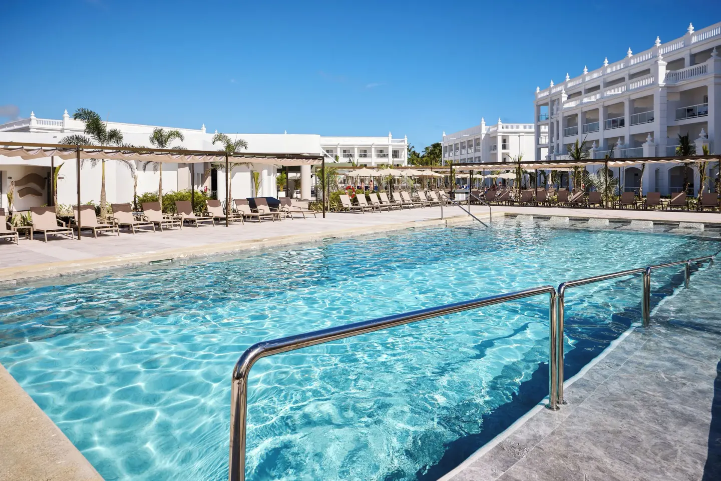 Large swimming pool with railings, loungers, and white buildings at Hotel Riu Palace Bavaro.