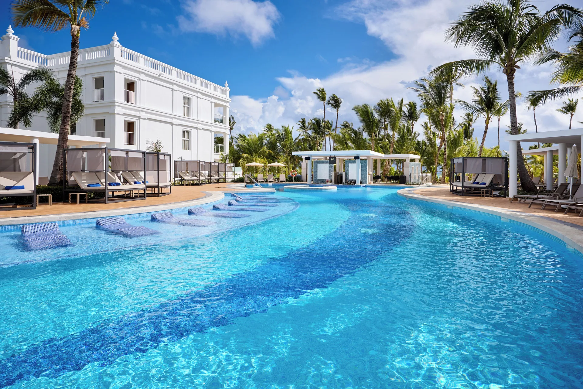 Curved pool with in-water loungers and cabanas beside palms at Hotel Riu Palace Bavaro.