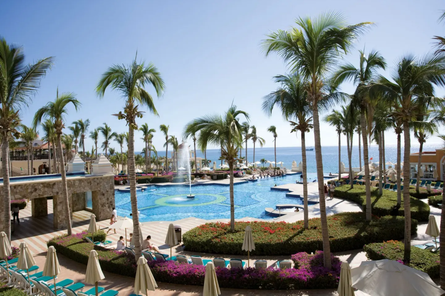 Palm-framed pool with a fountain and ocean beyond at Hotel Riu Palace Cabo San Lucas.