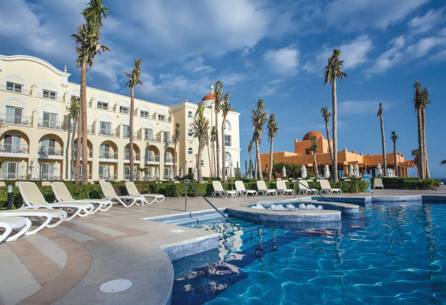 Curved pool edge, loungers, palms, and resort facade at Hotel Riu Palace Cabo San Lucas.