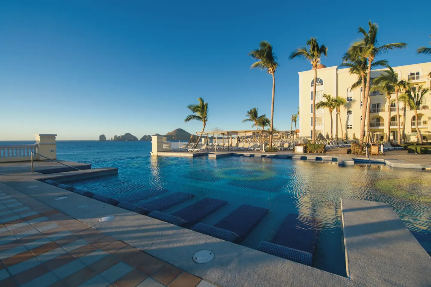Quiet pool terrace beside palms and resort buildings at Hotel Riu Palace Cabo San Lucas.