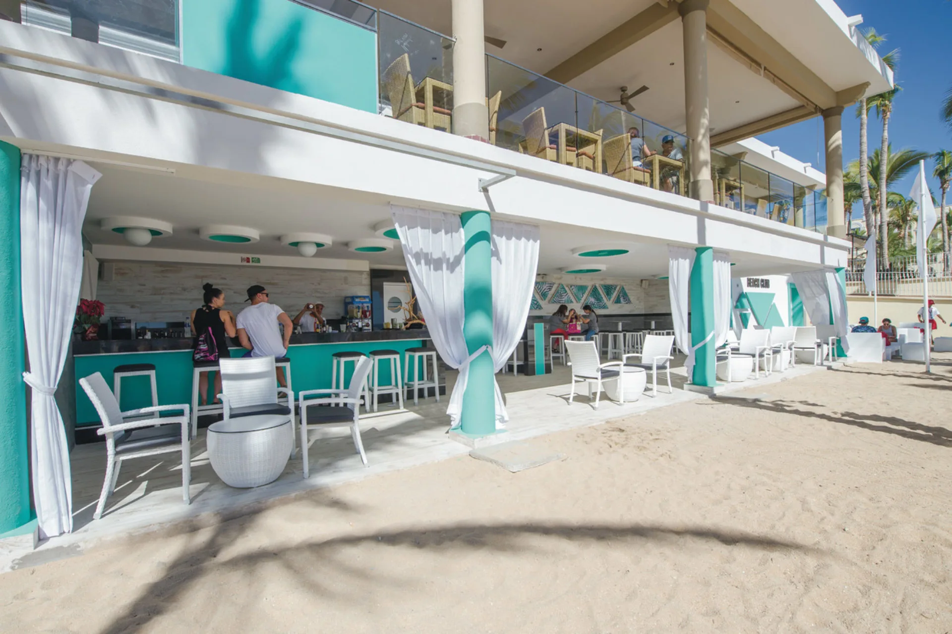 Beachfront bar with turquoise accents and sand-side seating at Hotel Riu Palace Cabo San Lucas.